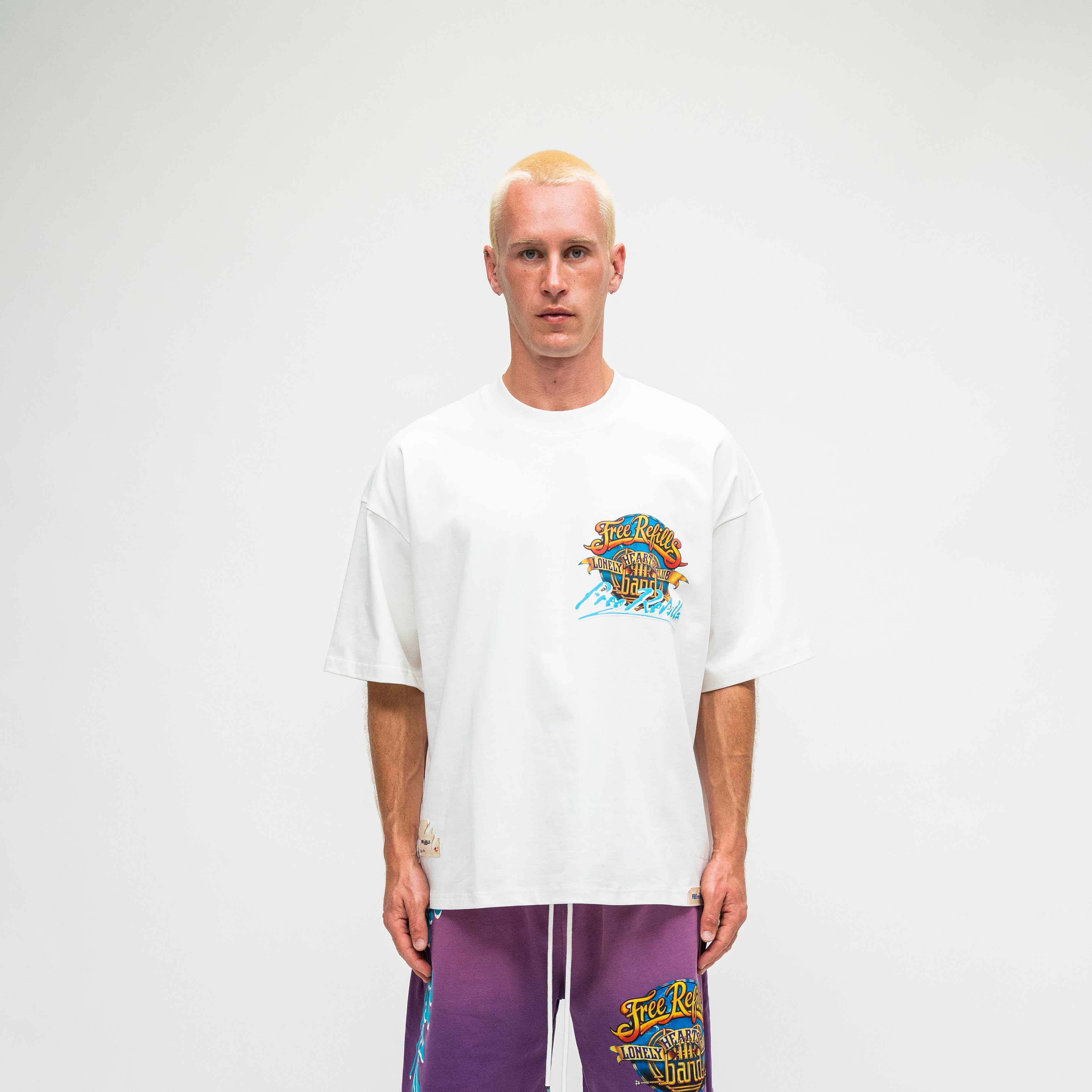 A man with a buzz cut stands against a light gray studio background wearing a white short-sleeve t-shirt with a colorful graphic on the left chest and purple drawstring sweatpants with a matching graphic.