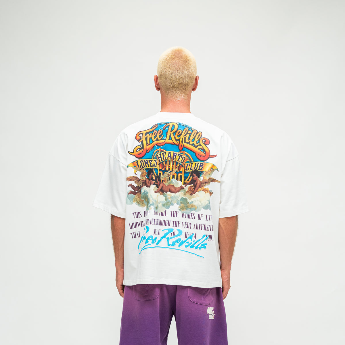 A man viewed from behind wearing a white graphic t-shirt with a large multicolor back print and purple sweatpants, set against a plain light gray studio background.