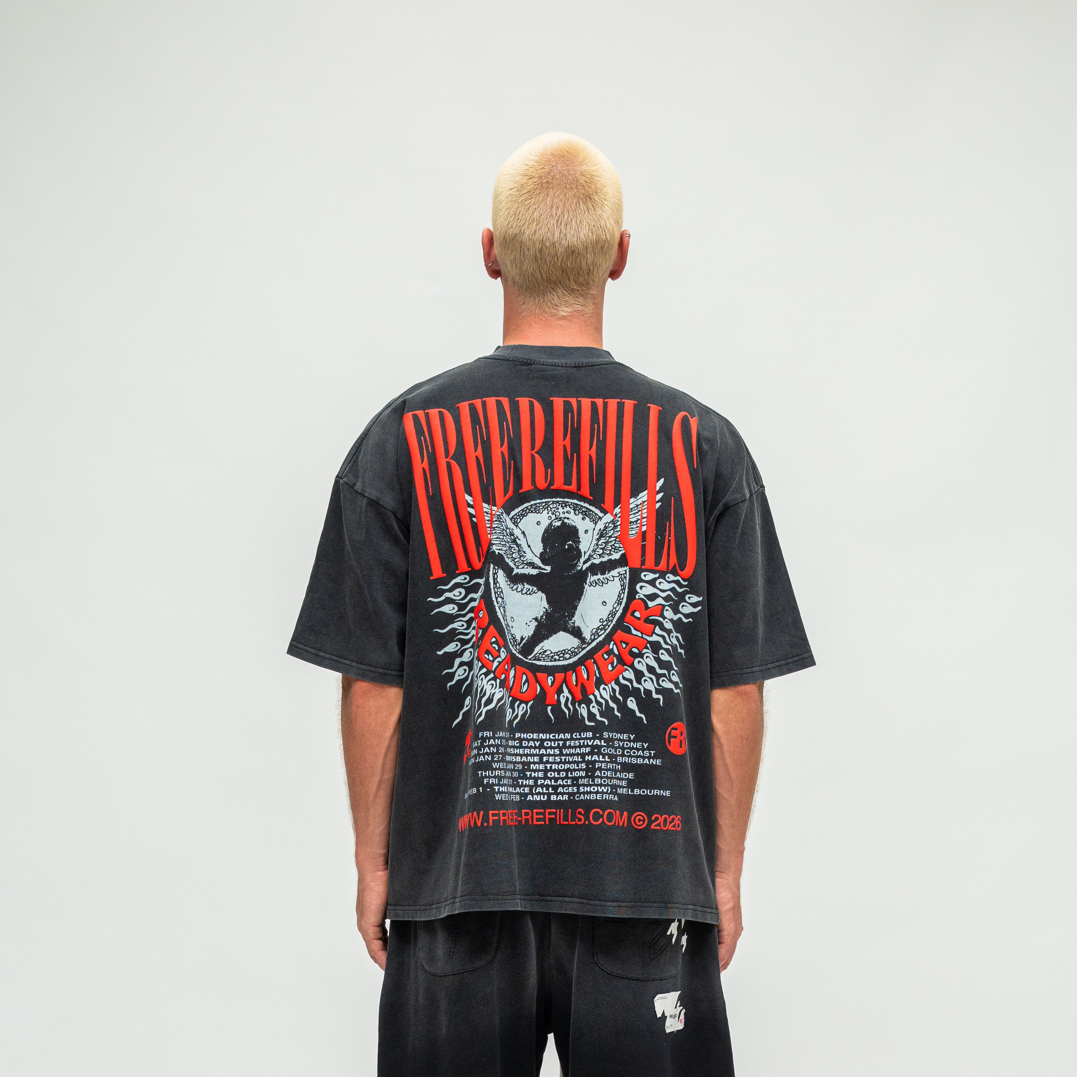 A man viewed from behind wearing a faded black graphic t-shirt with a large red and white 'FREE REFILLS' design on the back, paired with black pants, against a plain light gray background.
