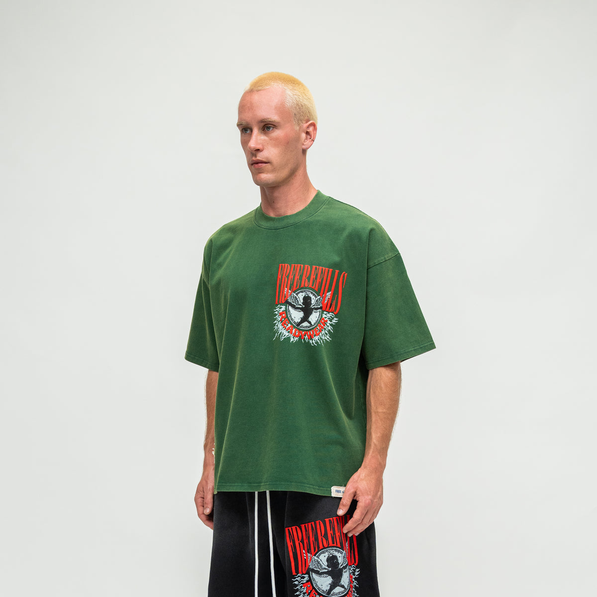 A man with bleached blonde hair wearing a green graphic t-shirt and black sweatpants with a matching graphic print, standing against a light gray studio background.