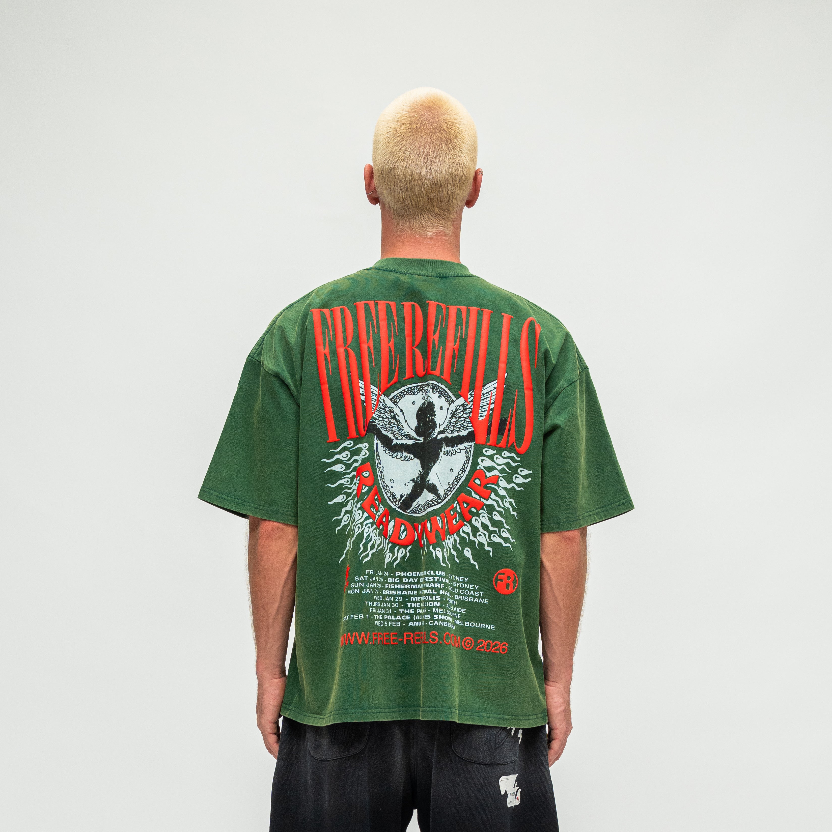 A man viewed from behind wearing a green graphic t-shirt with a large red, white, and black print on the back, paired with black pants, against a plain light gray background.