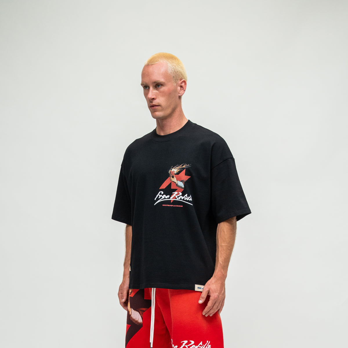 A man with blonde hair wearing a black t-shirt with a red and white graphic logo and red shorts, standing against a light gray background.