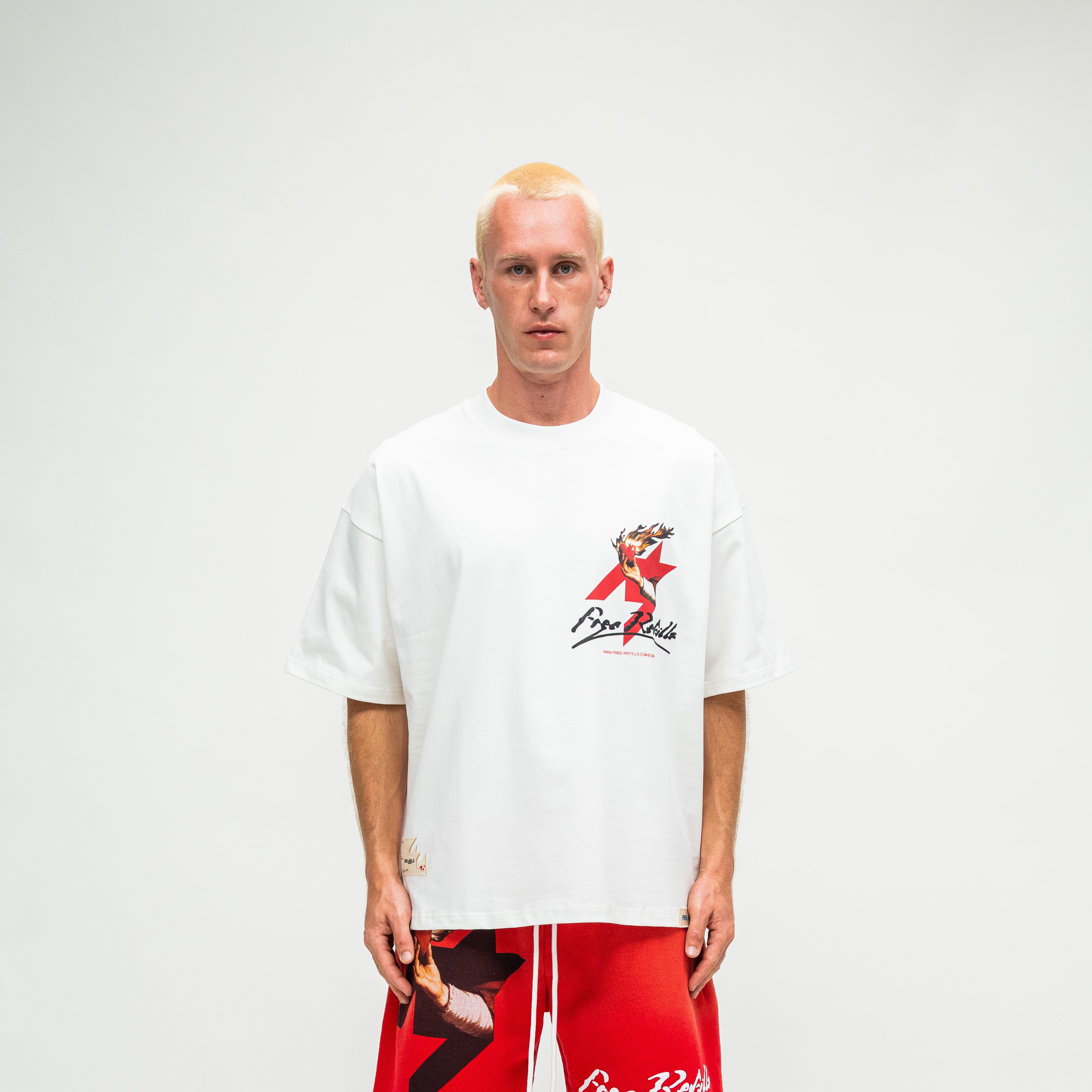 A man wearing an oversized white t-shirt with a red and black graphic on the left chest, paired with red shorts, standing against a plain light gray background.
