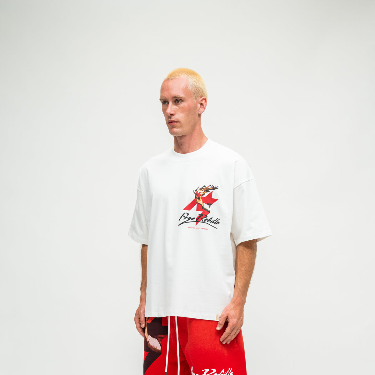 A man with platinum blonde hair wearing a white oversized t-shirt with a red and black graphic on the chest, paired with red shorts, standing against a plain light gray background.