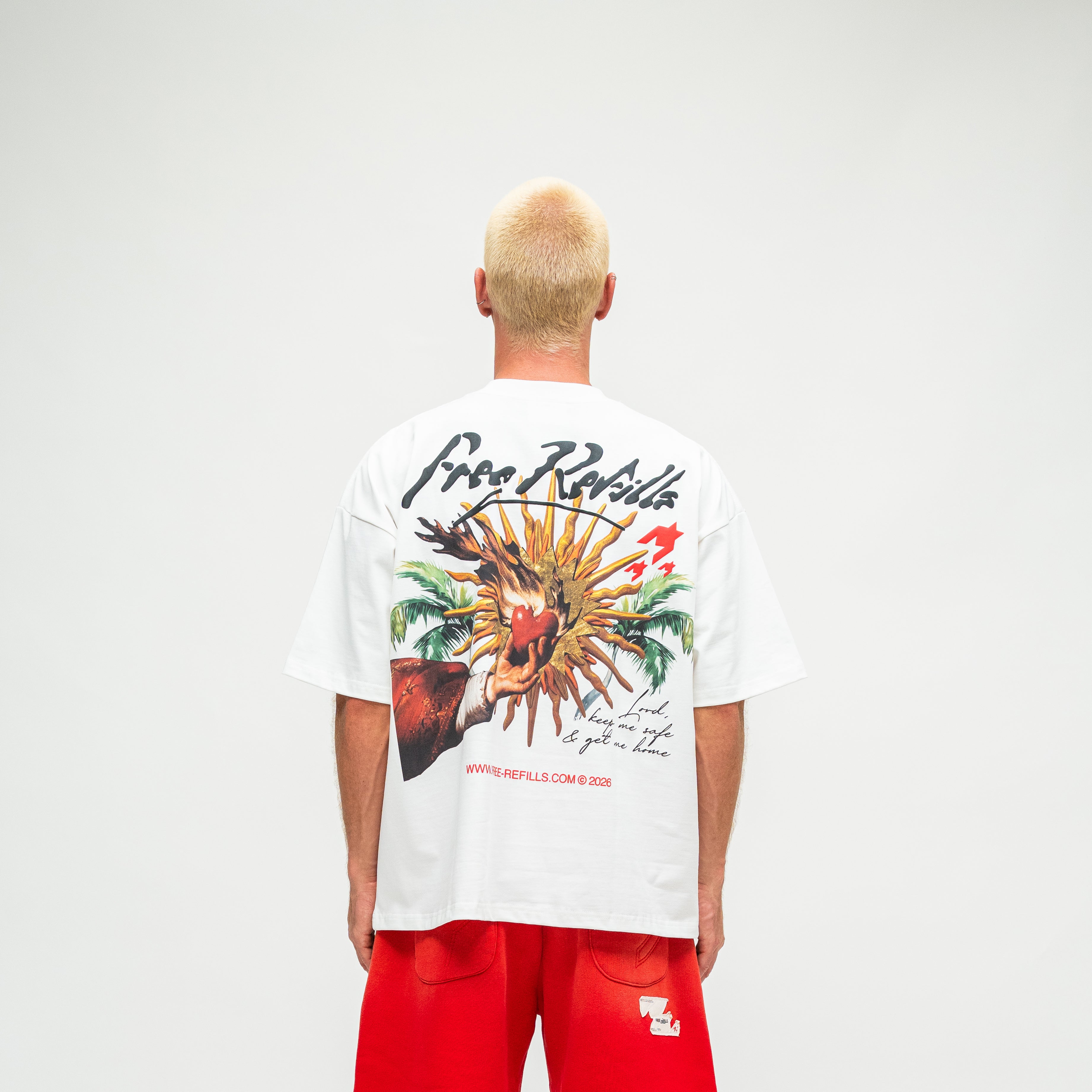 A man viewed from behind wearing a white t-shirt with a large colorful graphic print on the back and red shorts, standing against a light gray studio background.