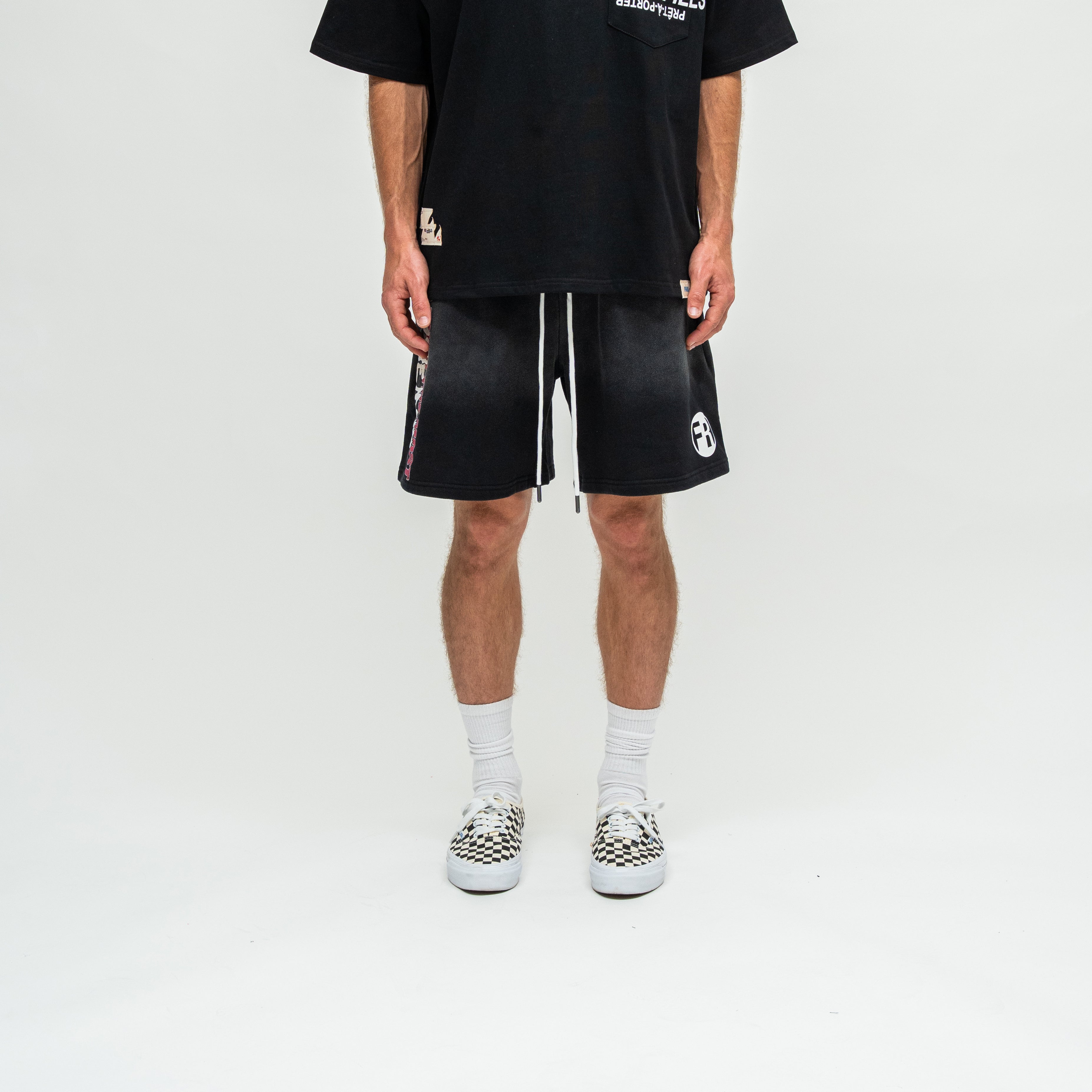 A model wearing black athletic shorts with white drawstrings, a black graphic t-shirt, white socks, and black-and-white patterned slip-on shoes, shown from the waist down against a white studio background.
