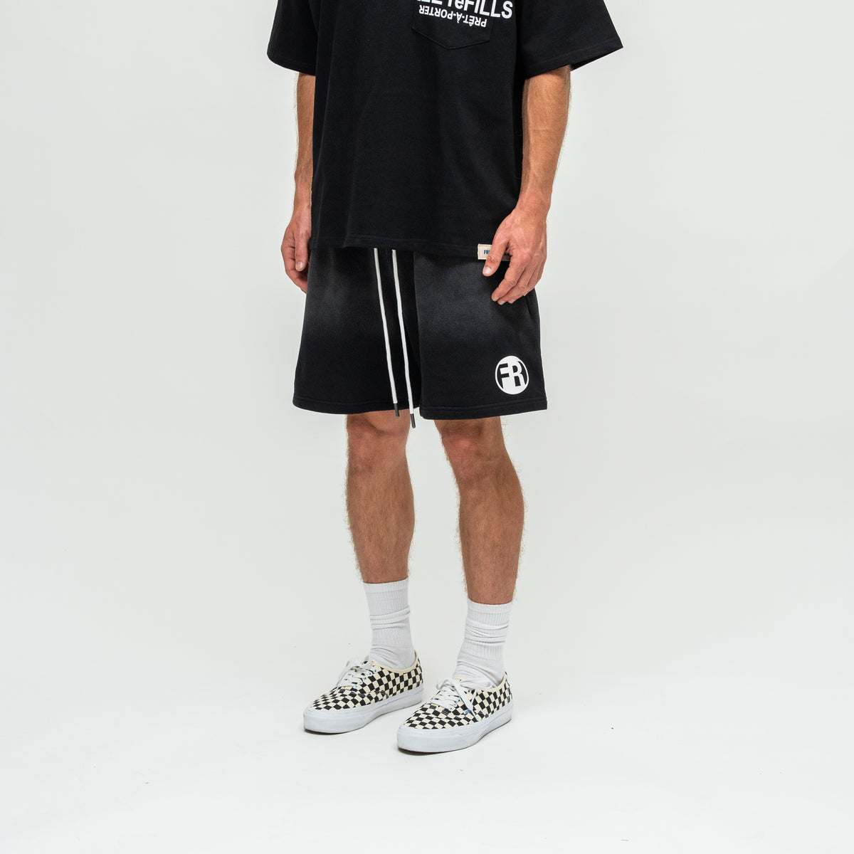A model wearing black athletic shorts with white side stripes and a white circular logo, paired with a black graphic t-shirt, white socks, and black-and-white checkered Vans sneakers, shot against a plain white studio background.