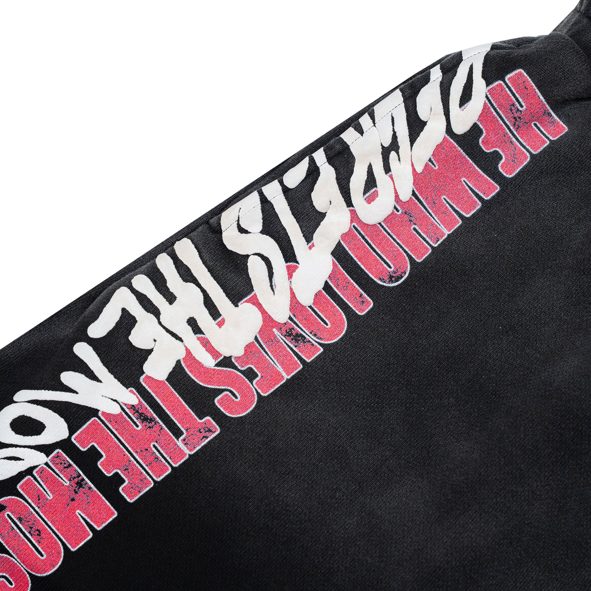 Close-up of a black garment featuring a diagonal graphic print with distressed white text and bold pink text reading 'HE WHO LOVES THE MOST.