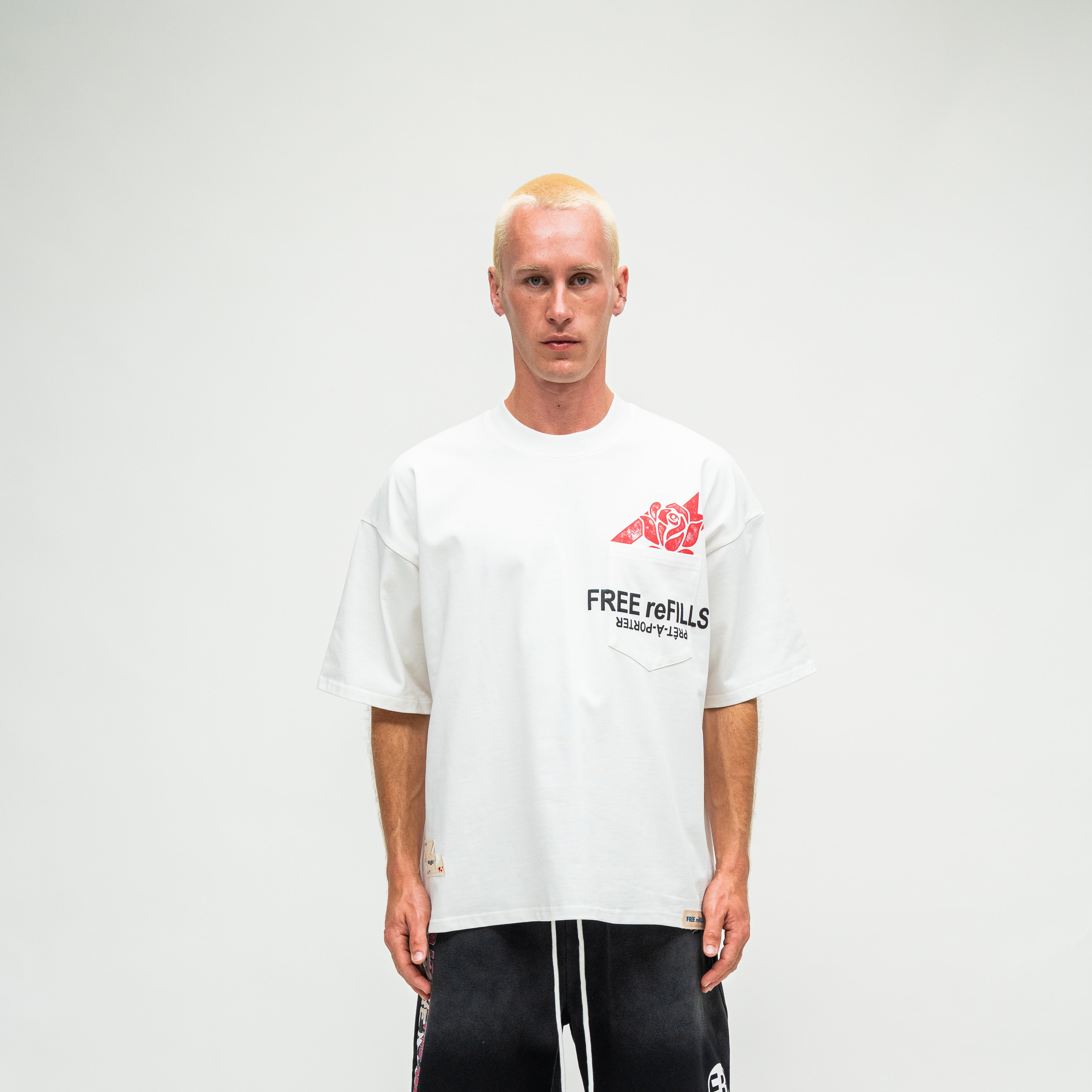 A man with platinum blonde hair stands against a light gray studio background wearing a white oversized t-shirt with a red graphic and black text on the chest, paired with black pants.