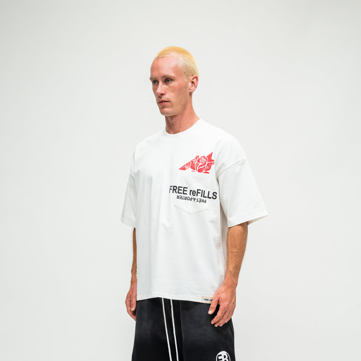 A man with blonde hair wearing a white t-shirt with a red fish graphic and black text that reads 'FREE REFILLS', paired with black shorts, standing against a light gray background.