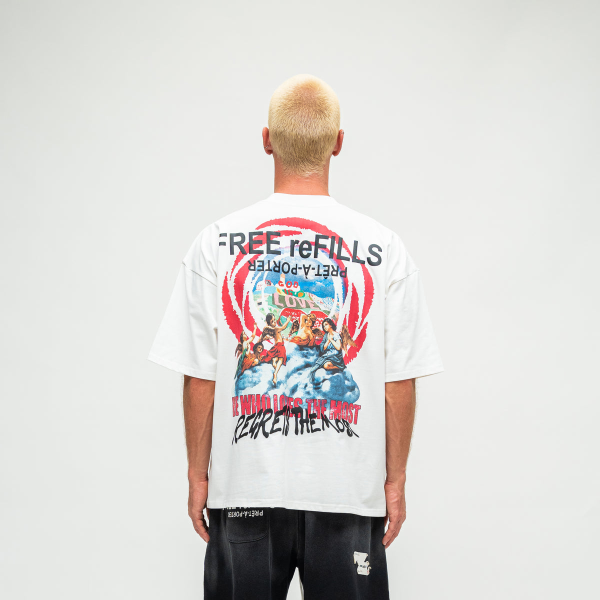 A man viewed from behind wearing a white graphic t-shirt with a large multicolor print on the back, paired with black shorts, against a plain light gray studio background.