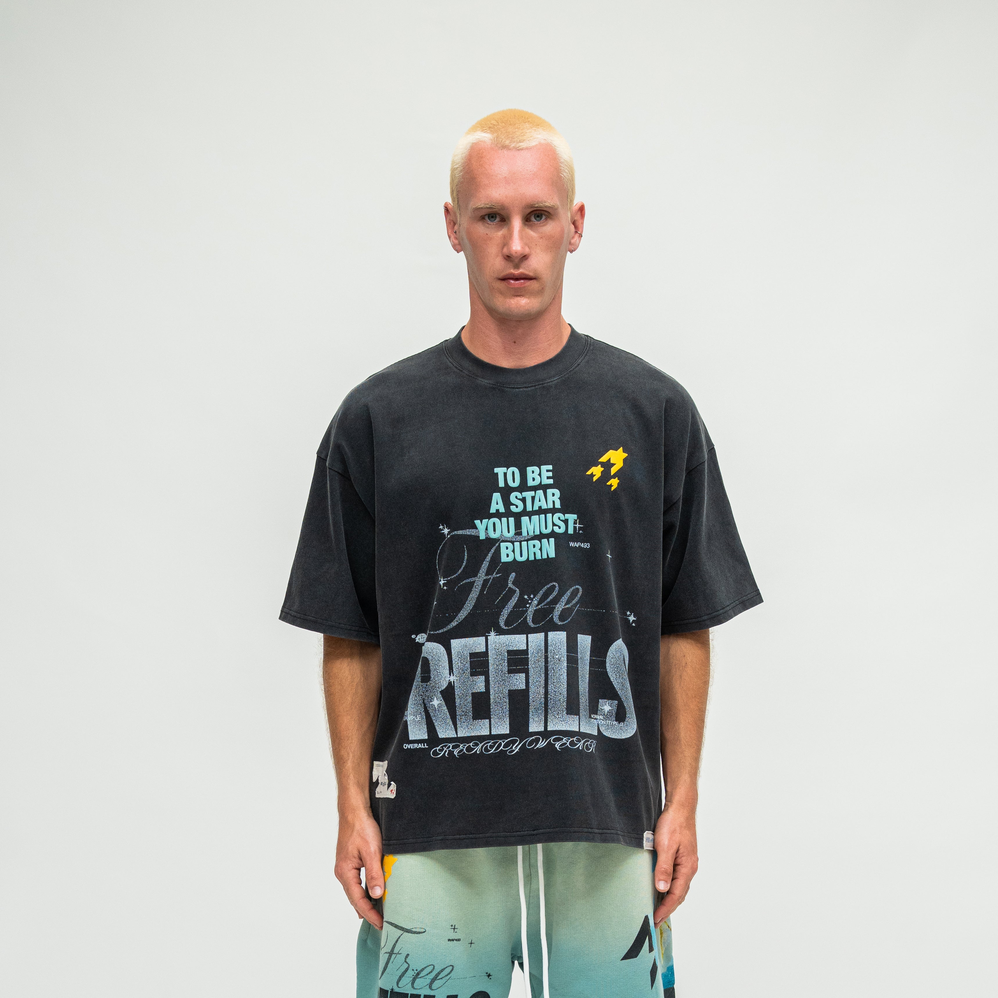 A man modeling a dark charcoal gray graphic t-shirt with 'Free REFILLS' and 'TO BE A STAR YOU MUST BURN' printed on the front, paired with light blue gradient shorts, against a plain light gray background.
