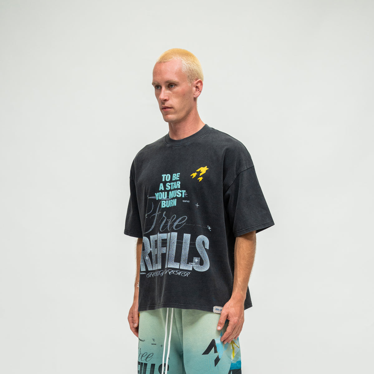 A man with bleached blonde hair stands against a light gray studio background wearing an oversized black graphic t-shirt and light blue gradient shorts. The t-shirt features a distressed yellow and teal graphic with the text 'TO BE A STAR YOU MUST BURN' and 'Free REFILLS'.