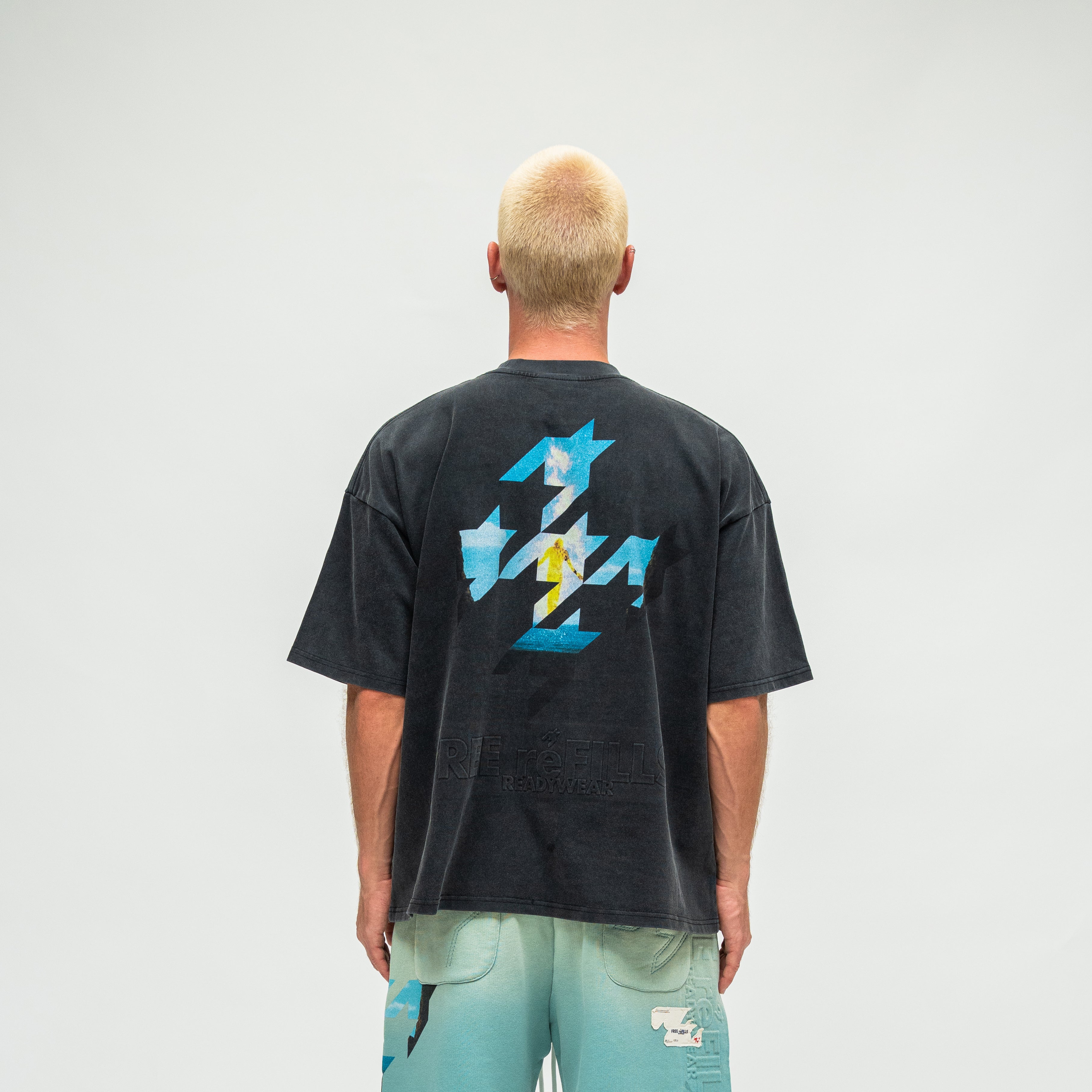 A man viewed from behind wearing a faded black graphic t-shirt with a large blue and white geometric design on the back, paired with light blue shorts, against a plain light gray background.
