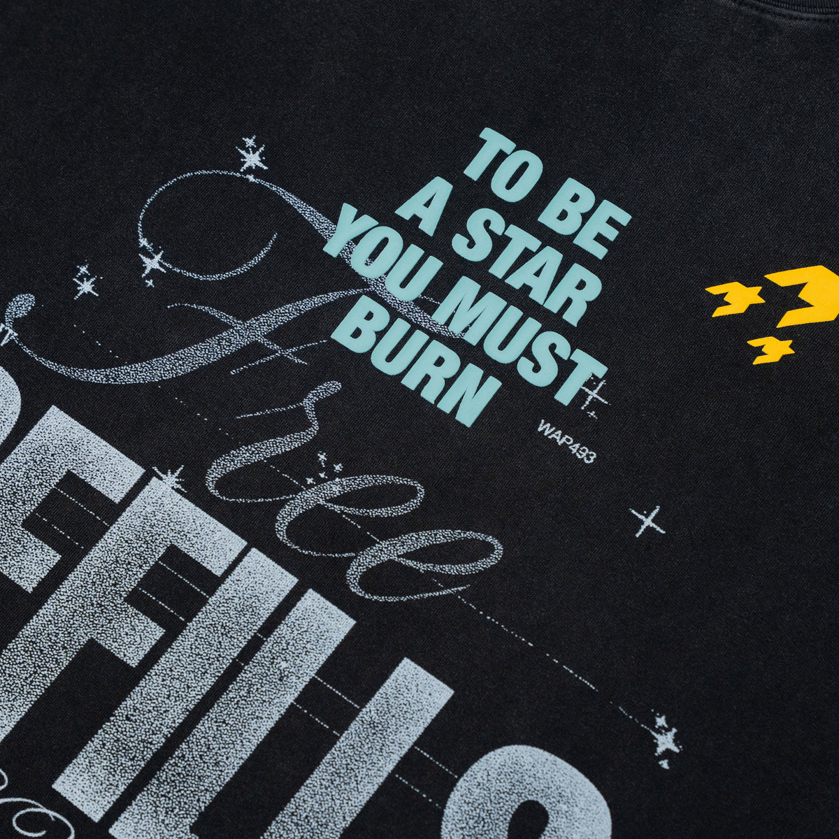 Close-up of a black graphic t-shirt featuring the text 'TO BE A STAR YOU MUST BURN' in light blue, distressed silver script, and a small yellow logo on the right side.