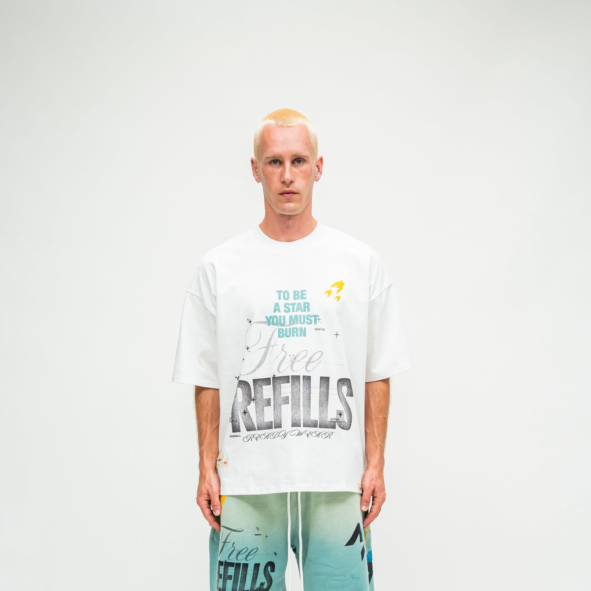 A man stands against a light gray studio background wearing an oversized white graphic T-shirt and teal shorts. The shirt has large gray and black text reading 'Free REFILLS' with smaller text above that says 'TO BE A STAR YOU MUST BURN' and a small yellow rocket graphic.