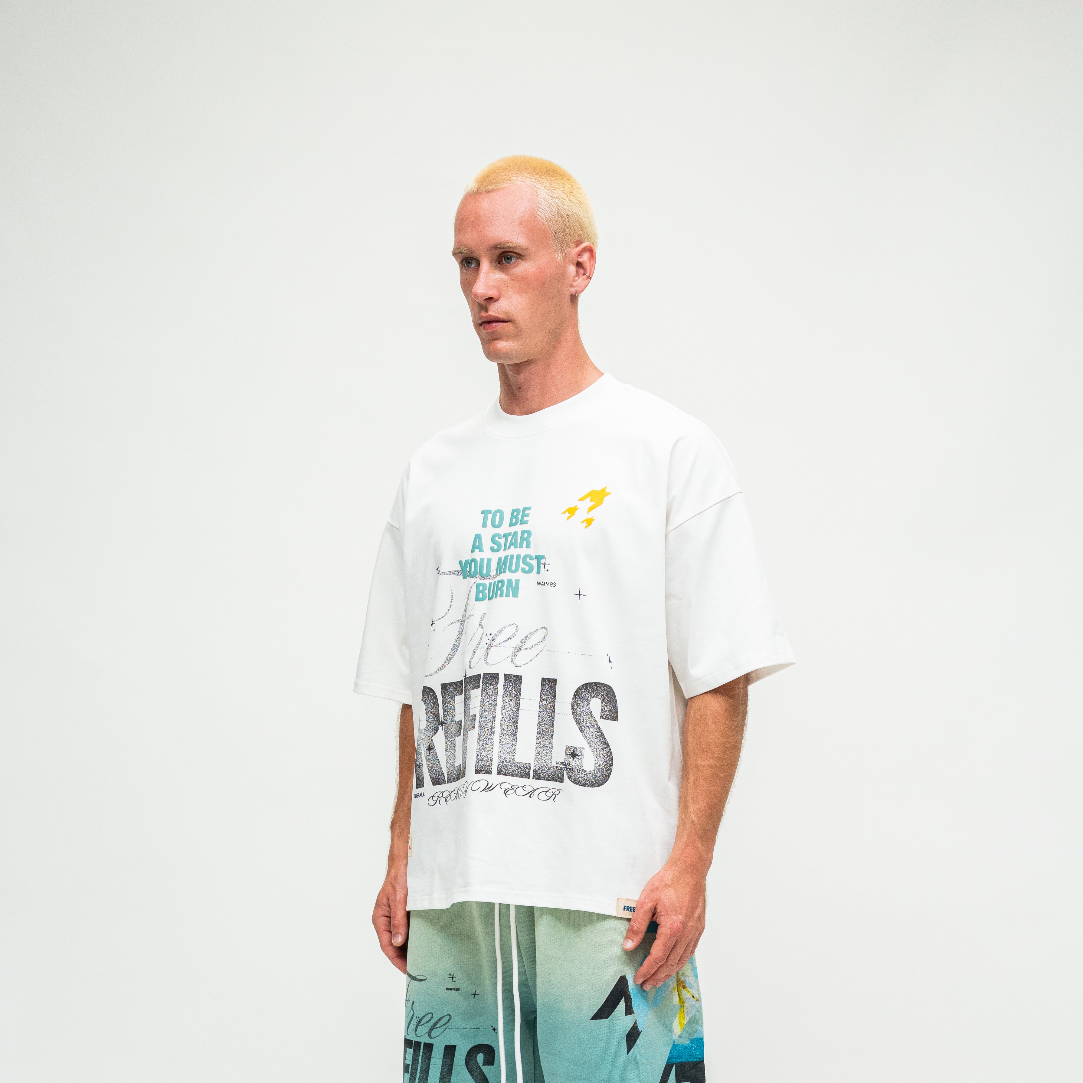A model with bleached blonde hair wearing an oversized white graphic t-shirt and teal shorts, standing against a plain white studio background.