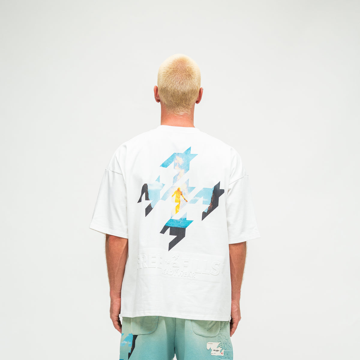 A man viewed from behind wearing a white t-shirt with a large graphic print on the back and light blue shorts, set against a plain light gray studio background.