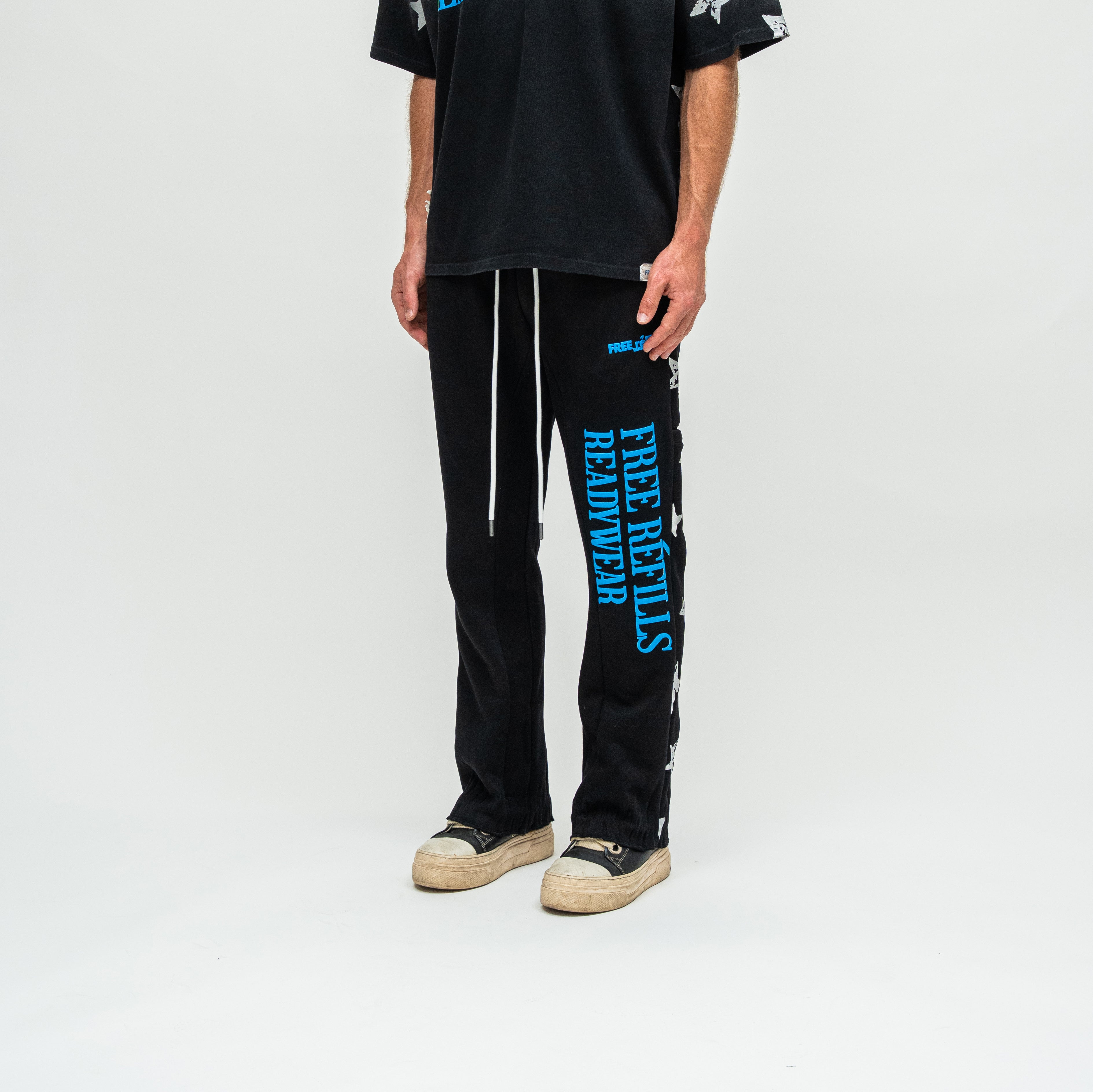 A model wearing black sweatpants with bright blue vertical text reading 'FREE REFILLS READYWEAR' down the left leg, paired with a black t-shirt and beige sneakers, against a plain white studio background.