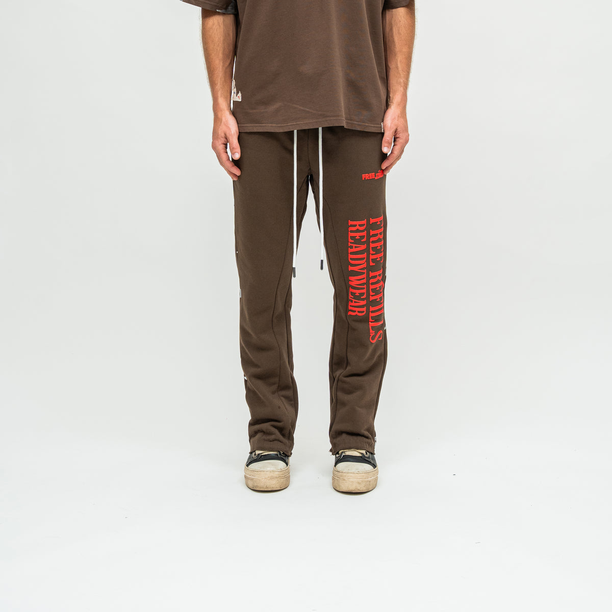 A person wearing brown sweatpants with red vertical 'READYWEAR' text on one leg, a matching brown T-shirt, and beige sneakers, photographed against a clean light gray studio background.