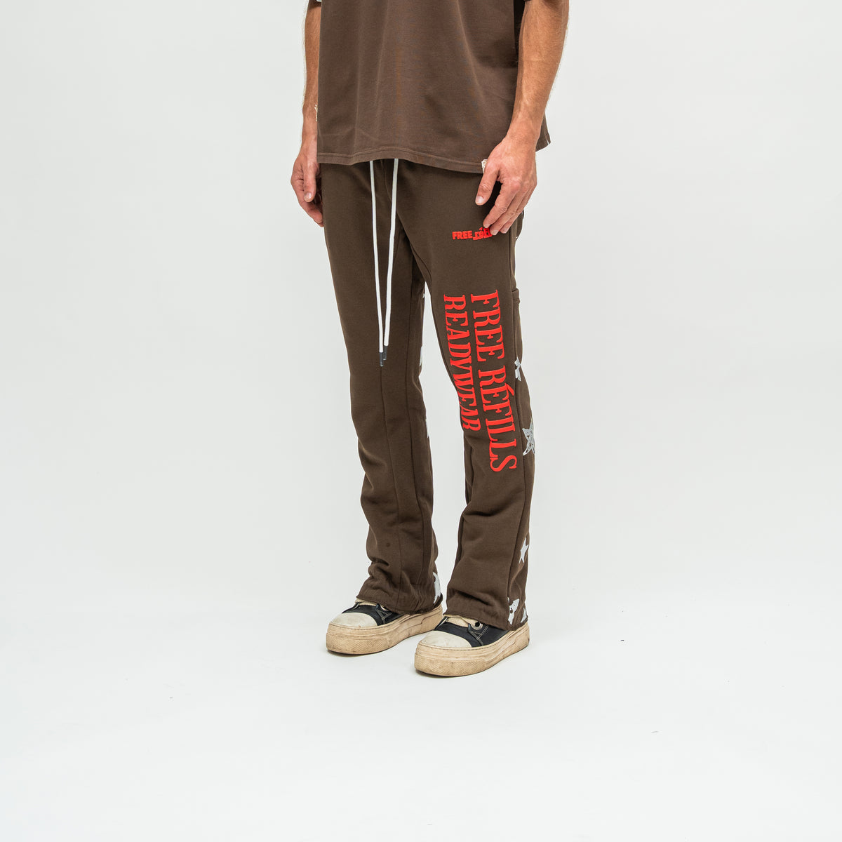 A person wearing brown sweatpants with bold red vertical text and a small red logo on the thigh, paired with a brown t-shirt and black-and-beige sneakers, photographed against a light gray studio background.