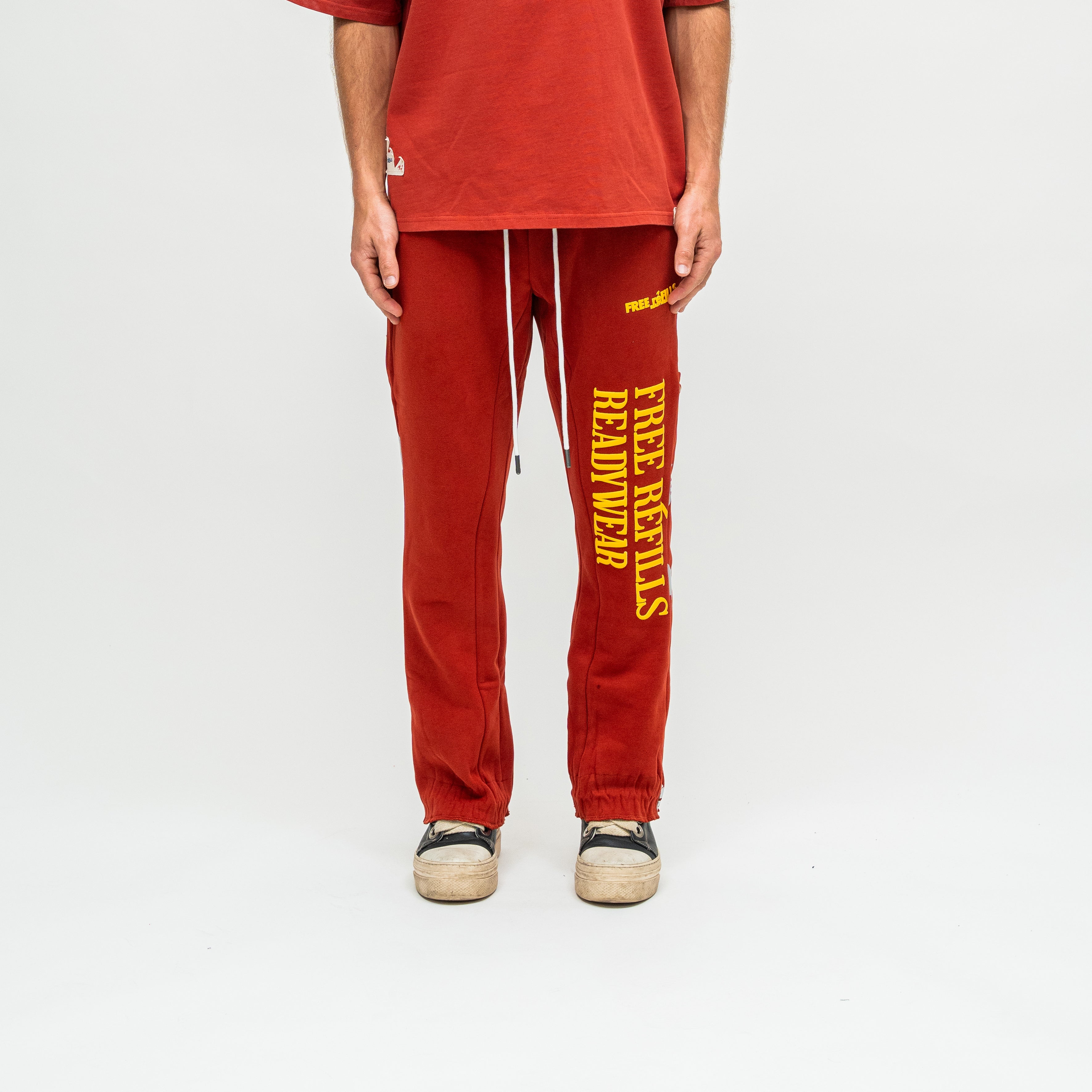 A person wearing red sweatpants with bold yellow vertical text reading 'FREE REFILLS READYWEAR' on the left leg, paired with a matching red t-shirt and gray sneakers, photographed against a plain light gray studio background.