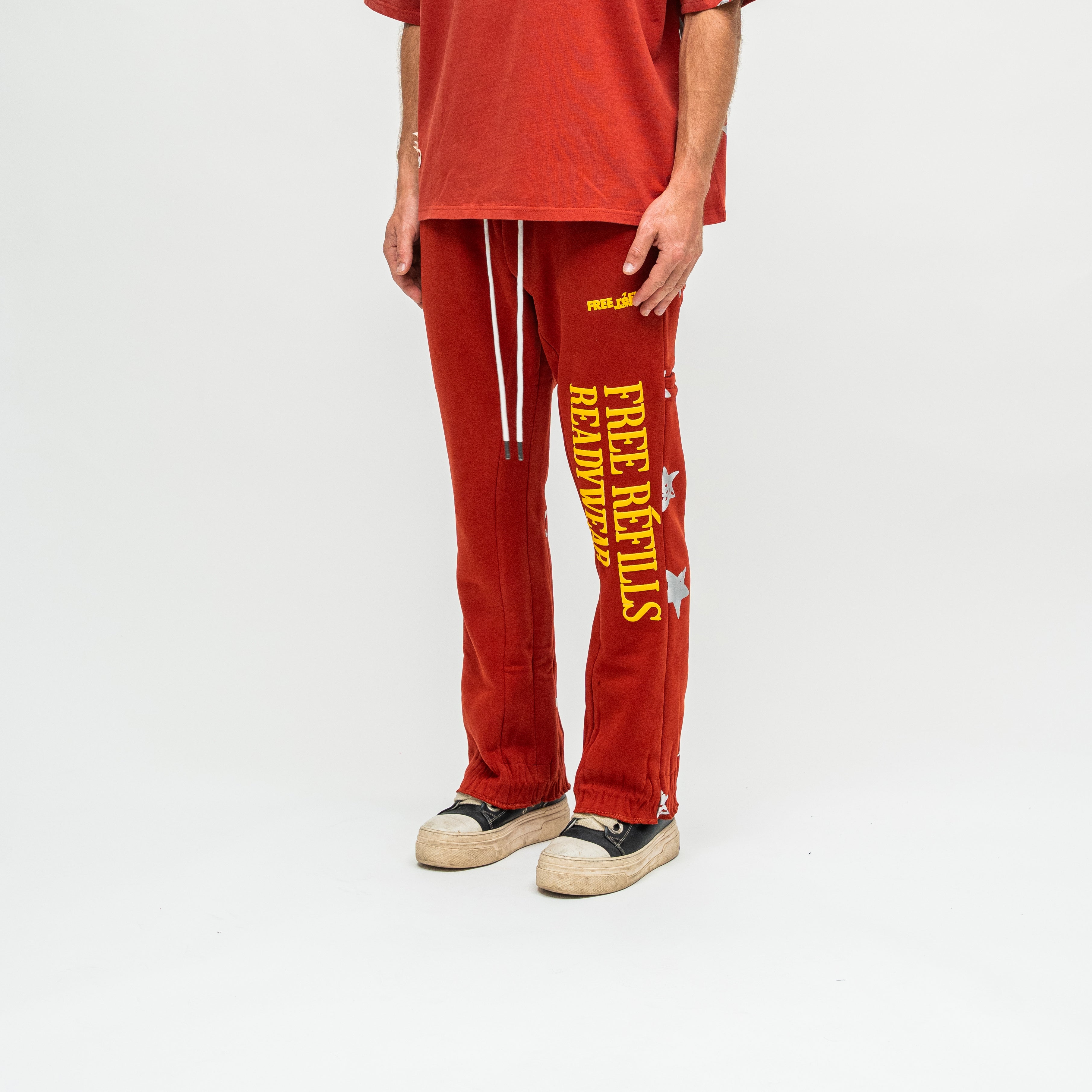A person wearing red sweatpants with bold yellow 'FREE REFiLLS READYWEAR' text and white drawstrings, paired with a matching red top and black-and-beige sneakers, photographed against a clean white studio background.