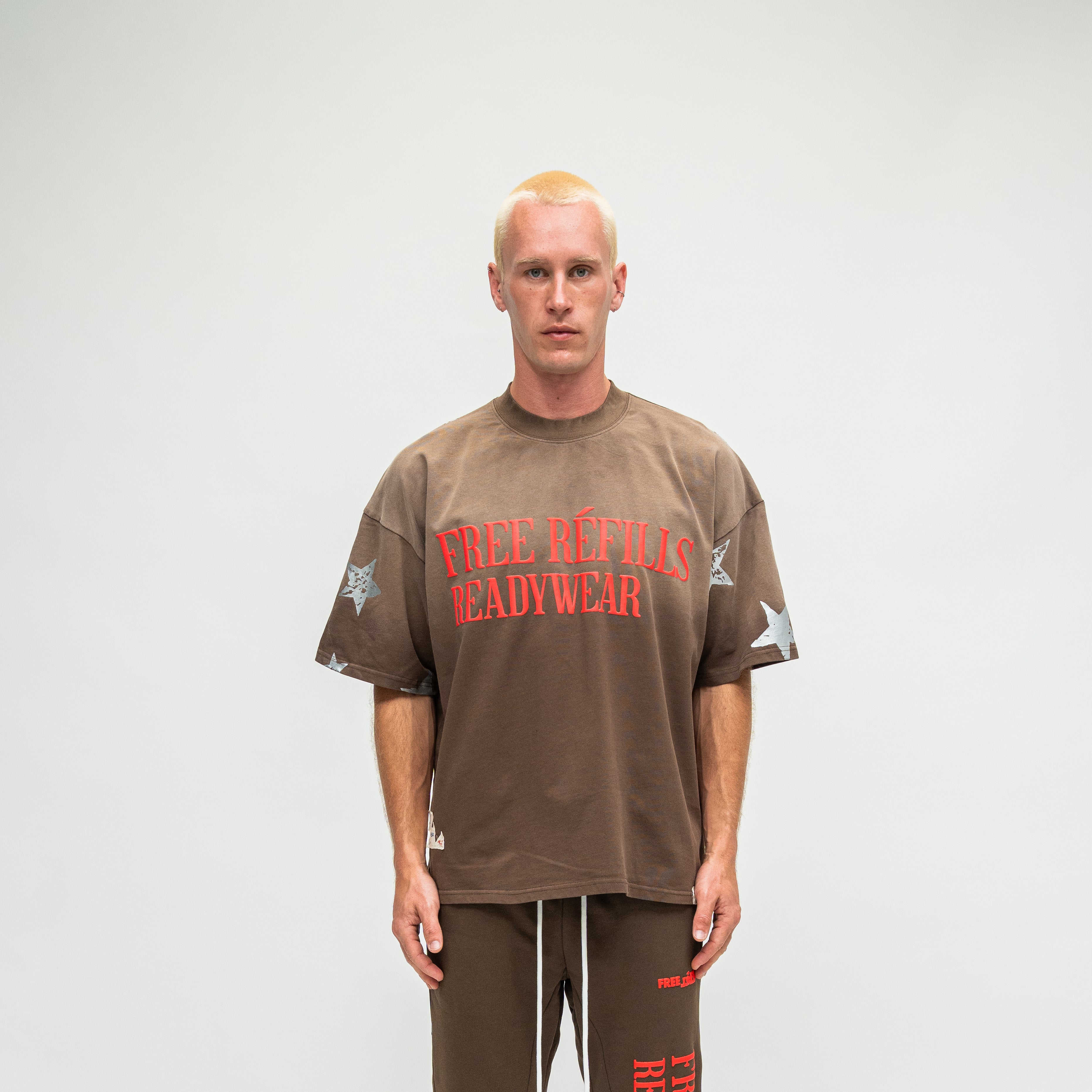 A man stands against a light gray studio background wearing an oversized brown t-shirt with red text reading 'FREE REFILLS READYWEAR' and white star graphics on the sleeves, paired with matching brown pants.