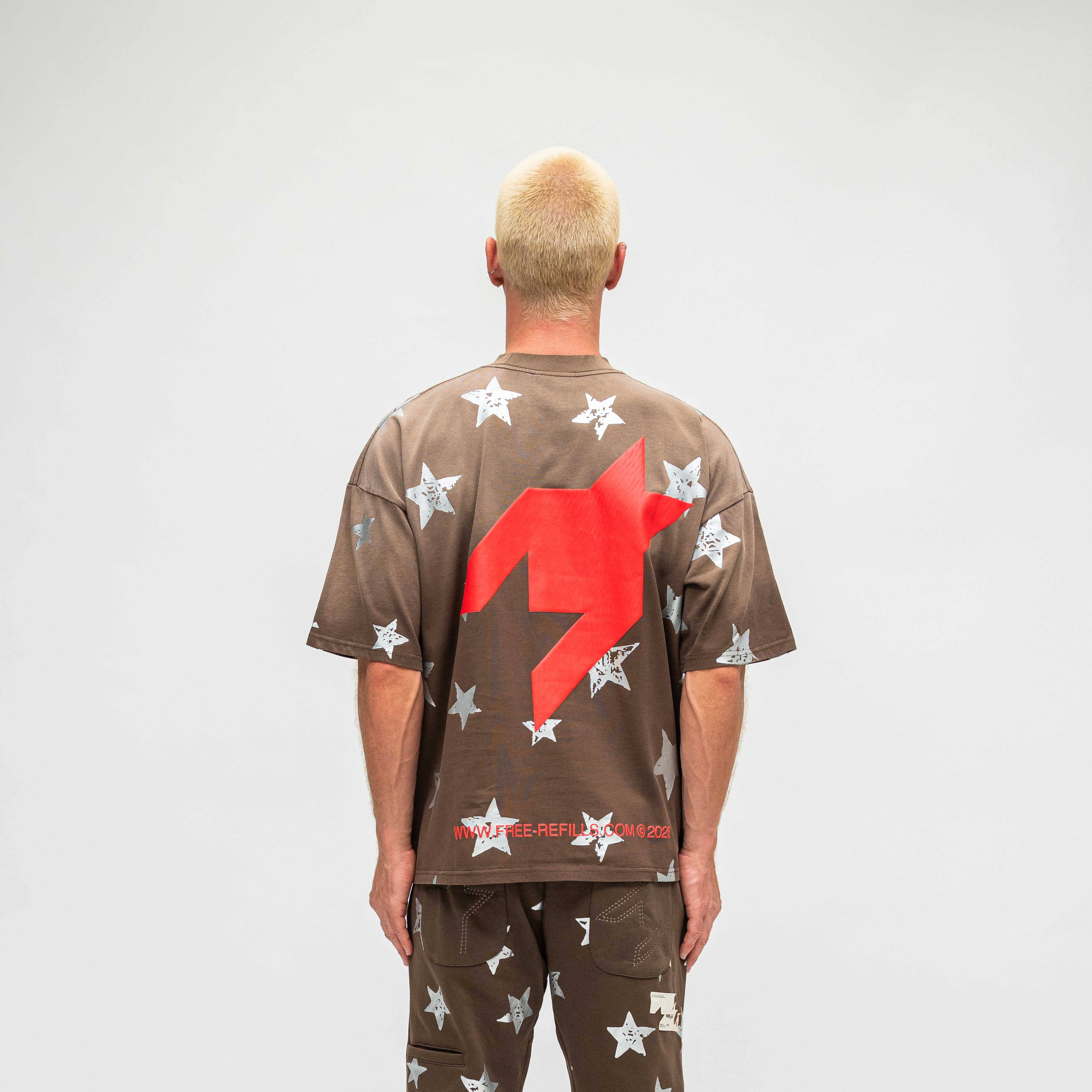 A man viewed from behind wearing a brown oversized t-shirt and matching pants, both covered in white star prints, with a large red graphic on the back of the shirt.