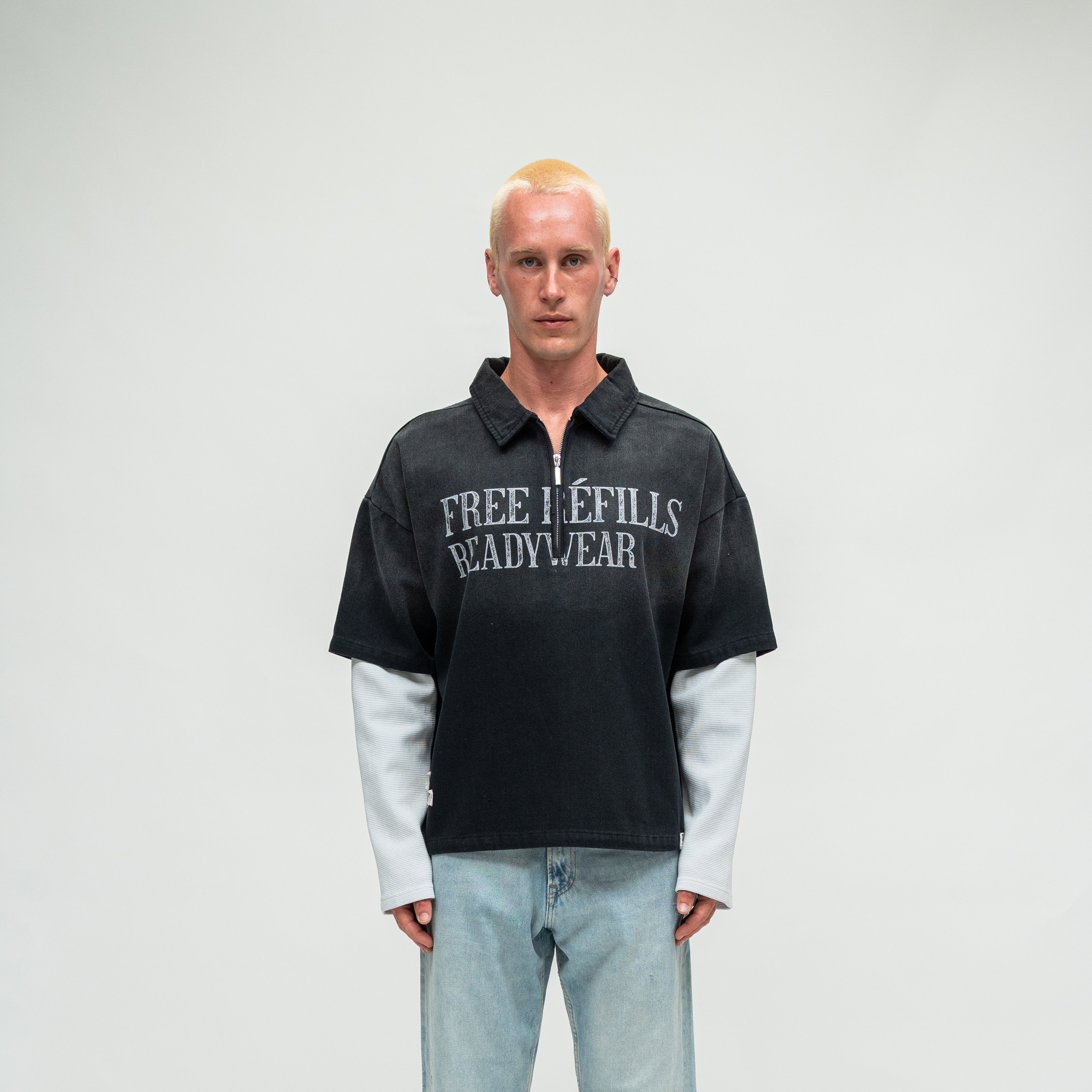 A model with short blonde hair stands against a light gray studio background wearing a black short-sleeve collared pullover with 'FREE REFILLS READYWEAR' printed in white across the chest, layered over a light gray long-sleeve shirt and paired with light blue jeans.