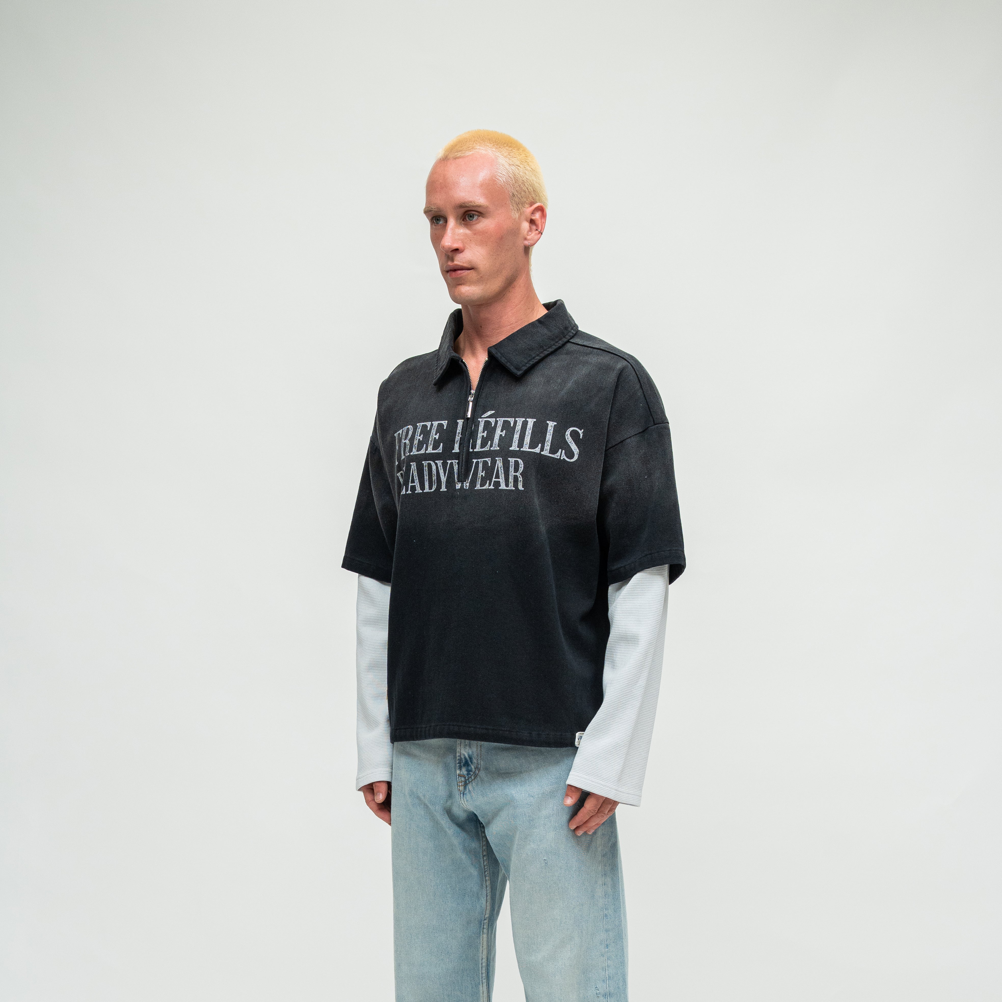 A model with bleached blonde hair stands against a light gray studio background wearing a black short-sleeve polo shirt with 'FREE REFILLS READYWEAR' printed in white across the chest, layered over a white long-sleeve shirt, and wearing light-wash blue jeans.