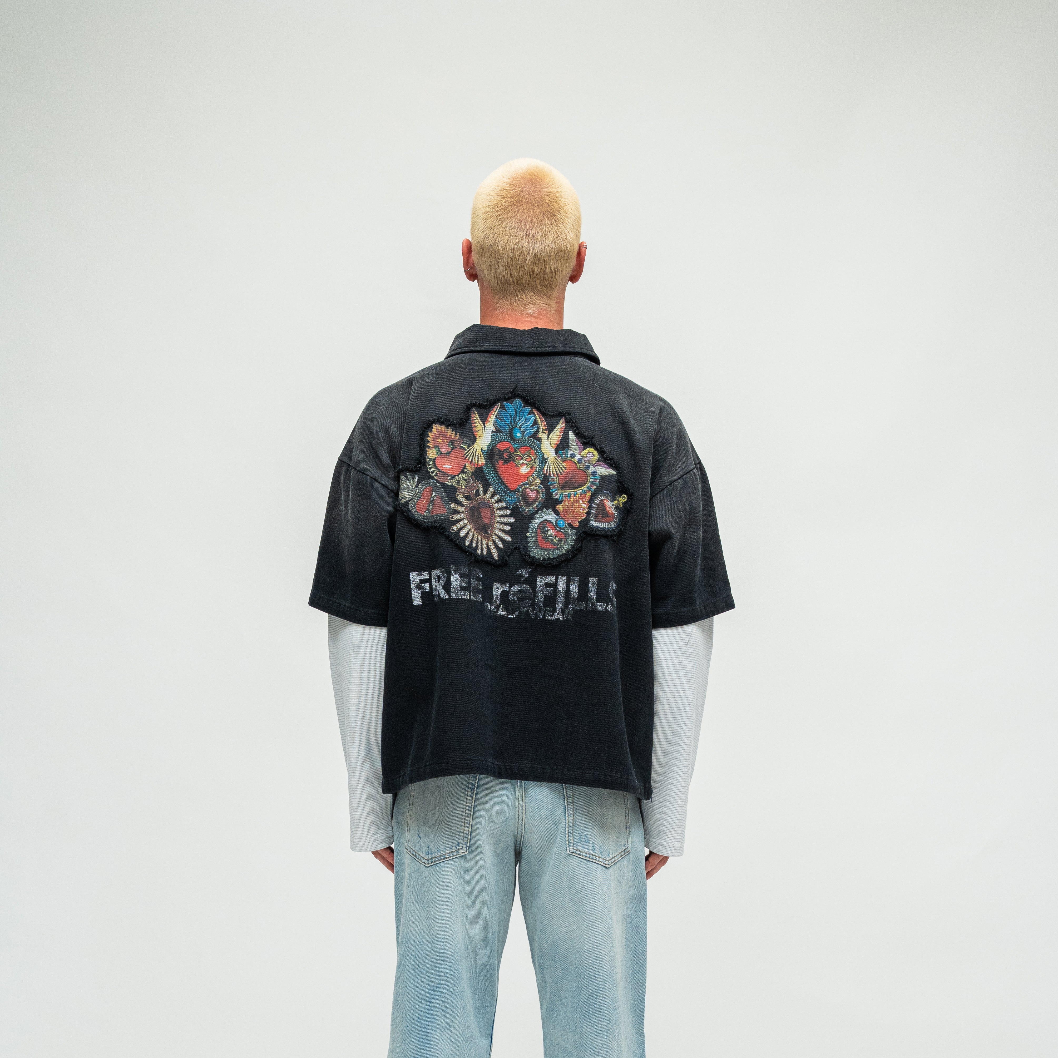 A model viewed from behind wearing a black short-sleeve button-up shirt with a large colorful graphic and 'FREE REFILLS' text on the back, layered over a white long-sleeve shirt and light blue jeans, against a light gray studio background.