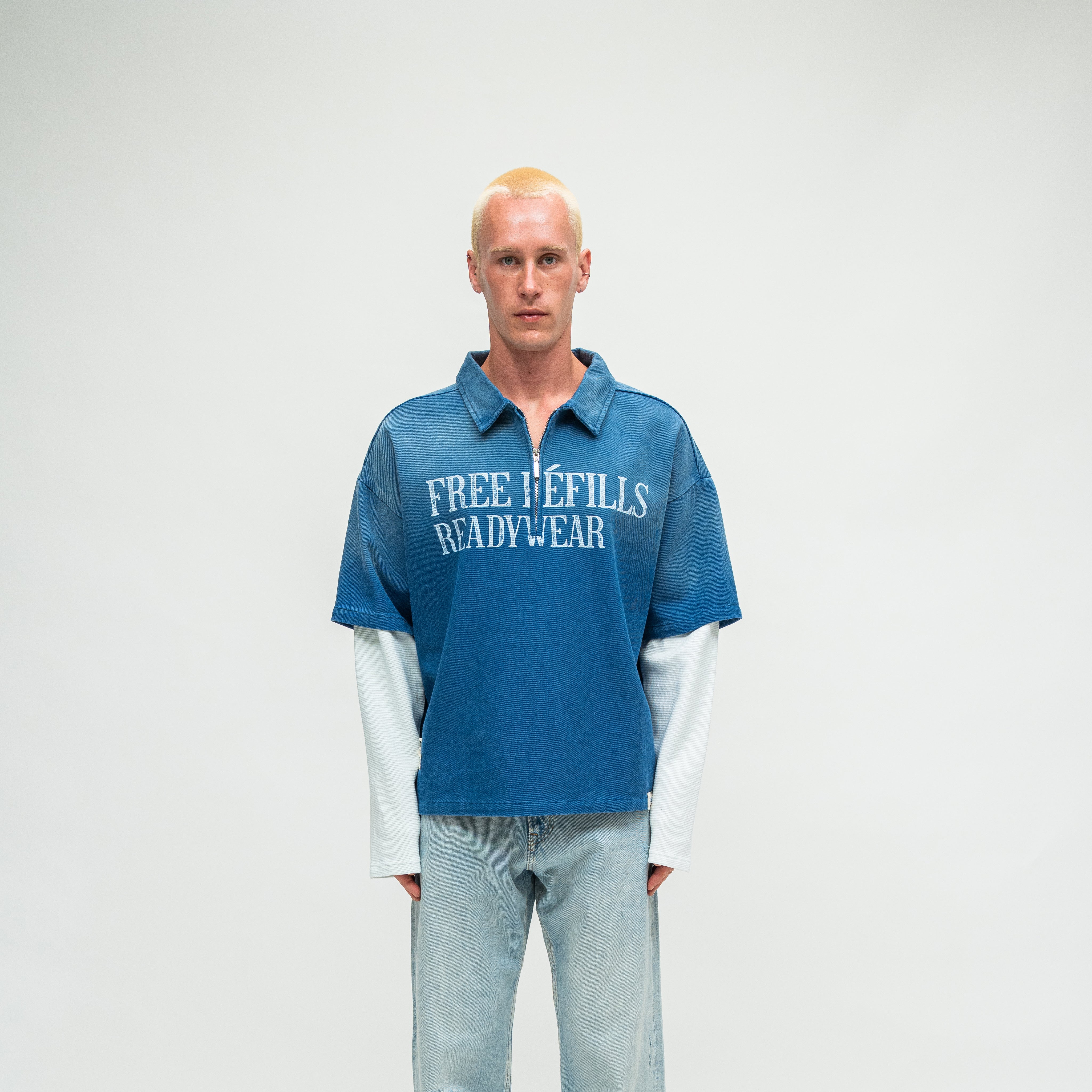 A man stands against a light gray studio background wearing a blue short-sleeve polo-style shirt with a white front text overlay reading 'FREE REFILLS READYWEAR' and light blue jeans.