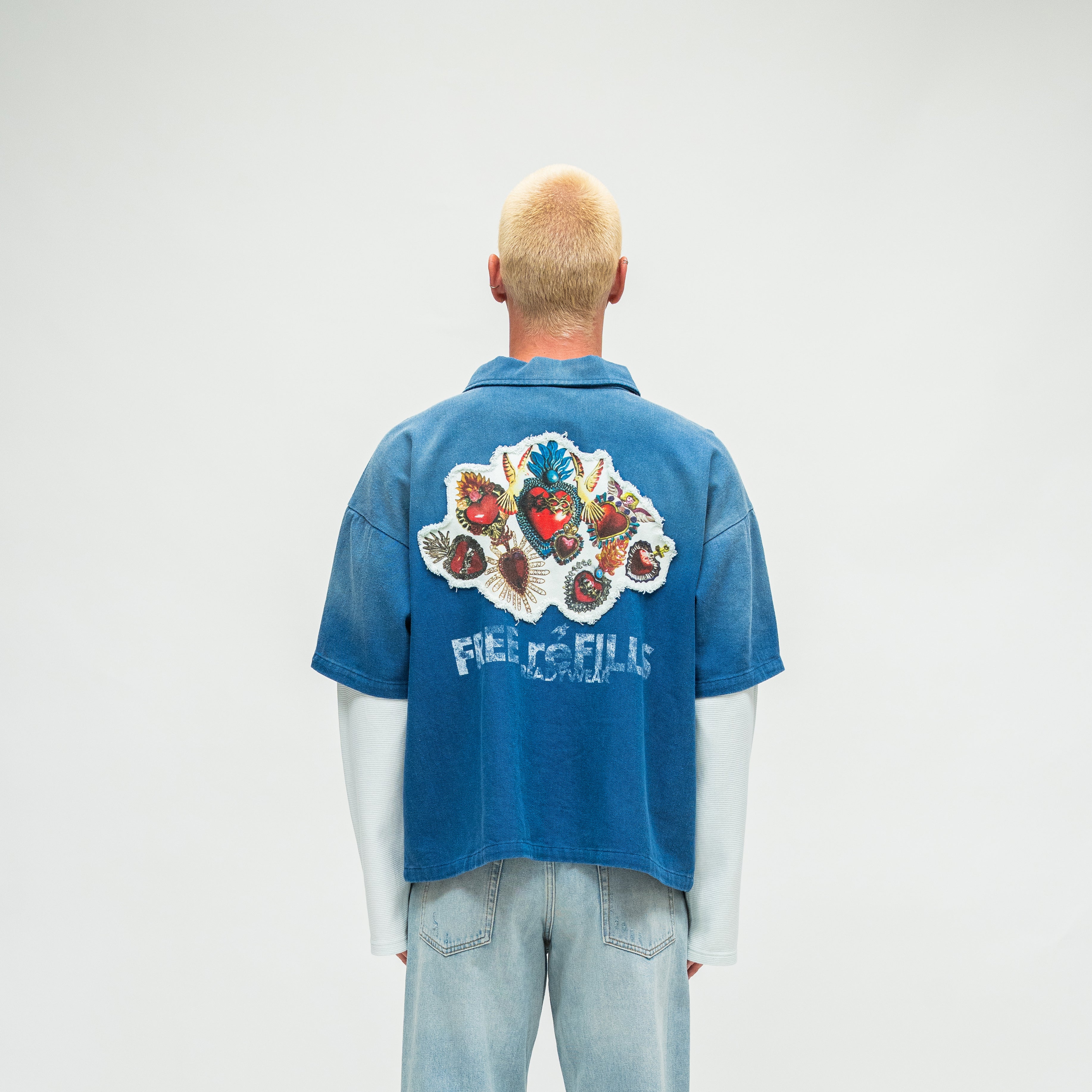 A man viewed from behind wearing a blue short-sleeve collared shirt with a large multicolored graphic and the word 'FREE REFILLS' across the back, paired with light blue jeans against a plain light gray studio background.