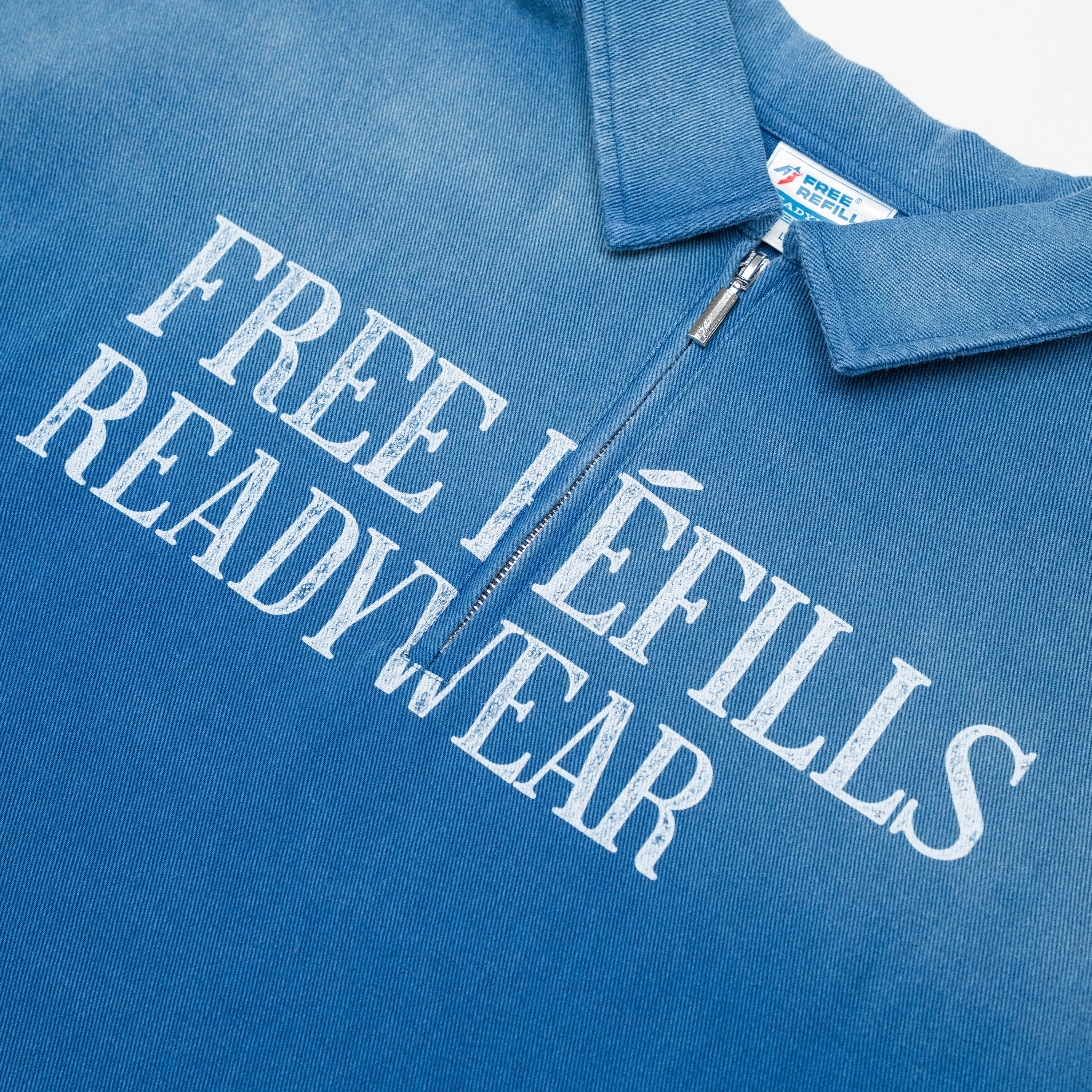 A close-up of a blue zip-up polo shirt with distressed white text reading 'FREE REFILLS READYWEAR' across the chest. The collar and a silver zipper are visible, along with a small neck label.